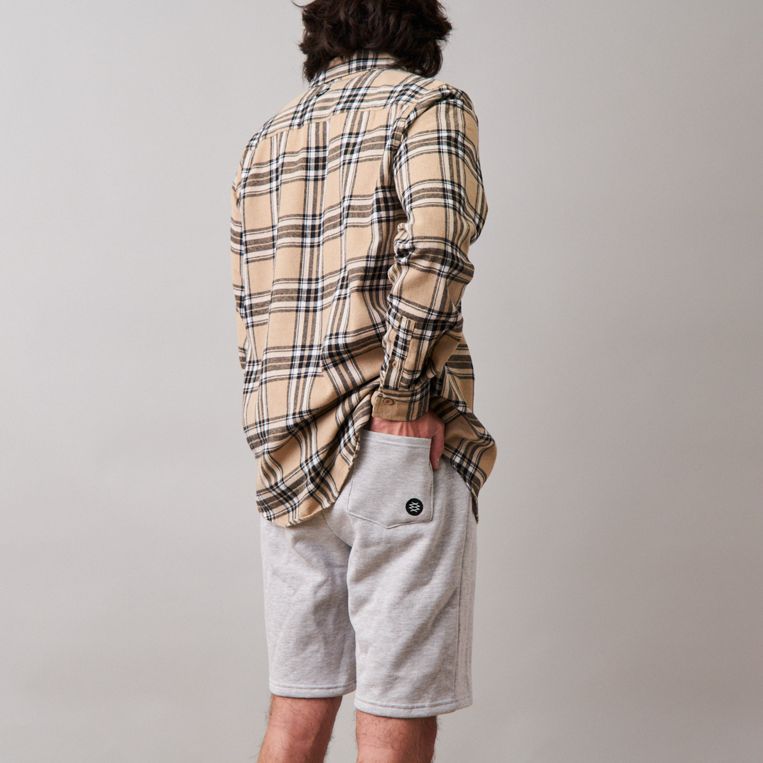 Man facing away from the camera wearing a beige long sleeve flannel and gray jogger shorts. The logo is visible on the lower corner of the back pocket.