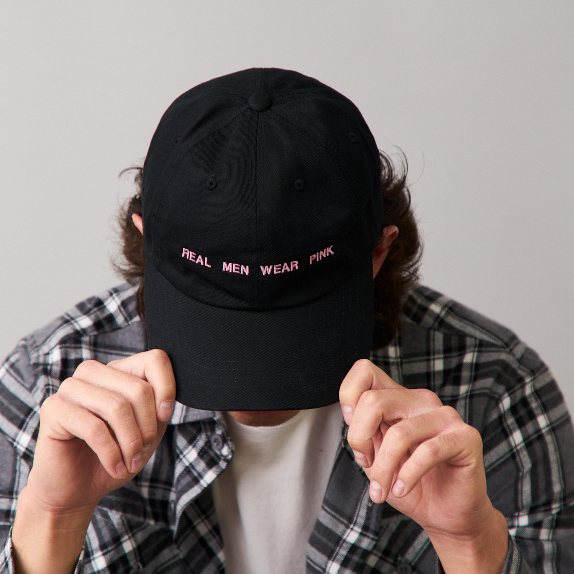 Man sitting forward with his head down, wearing a black dad cap with 'REAL MEN WEAR PINK' printed on the front, and a long sleeve gray flannel shirt.