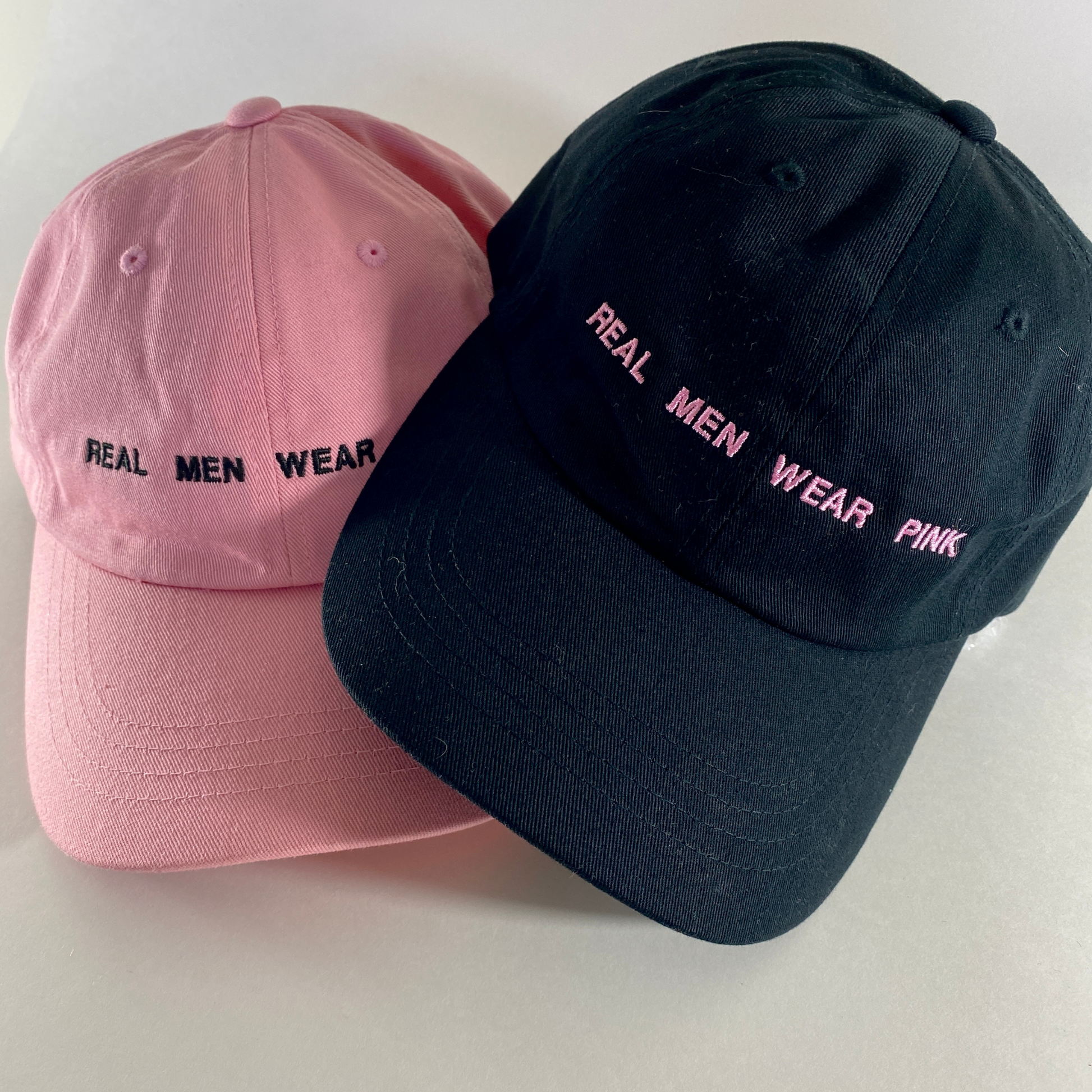 pink dad cap and black dad cap with REAL MEN WEAR PINK printed on front.