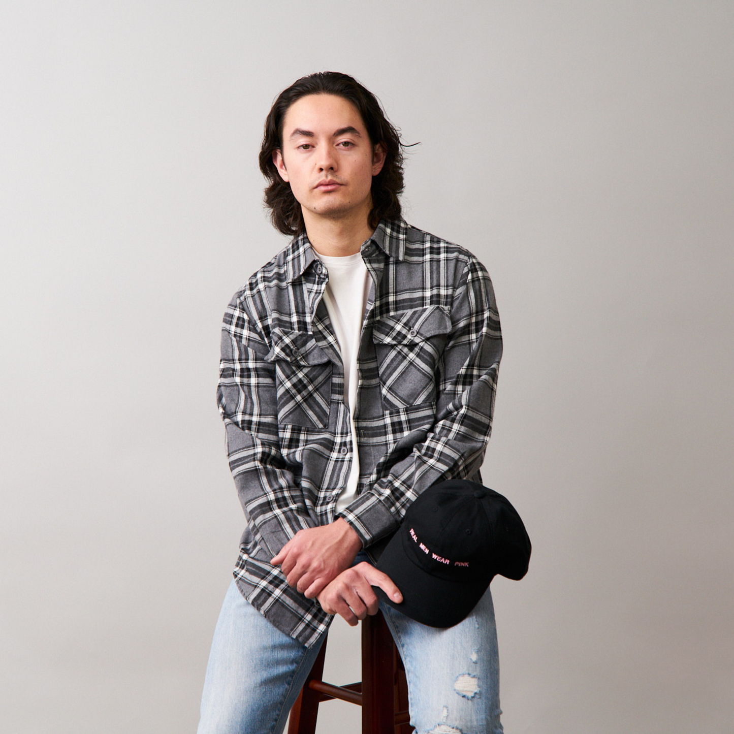 man sitting on a stool wearing long sleeve gray flannel, white tshirt, blue jeans and holding a black dad cap with REAL MEN WEAR PINK printed on front.