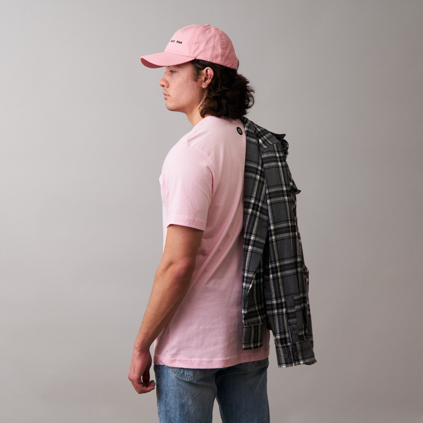 side view of man standing wearing pink tshirt with logo on the center backside and wearing blue jeans. He is wearing a pink dad cap with REAL MEN WEAR PINK printed on front, with a gray flannel shirt tossed over his shoulder.