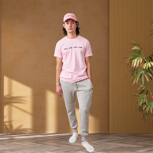 Man wearing gray fleece jogger sweatpants with ribbed bottom cuffs. The joggers have a tapered slim fit and a drawstring waistband, paired with a pink t-shirt and pink dad cap, both featuring 'REAL MEN WEAR PINK' printed on the front. 