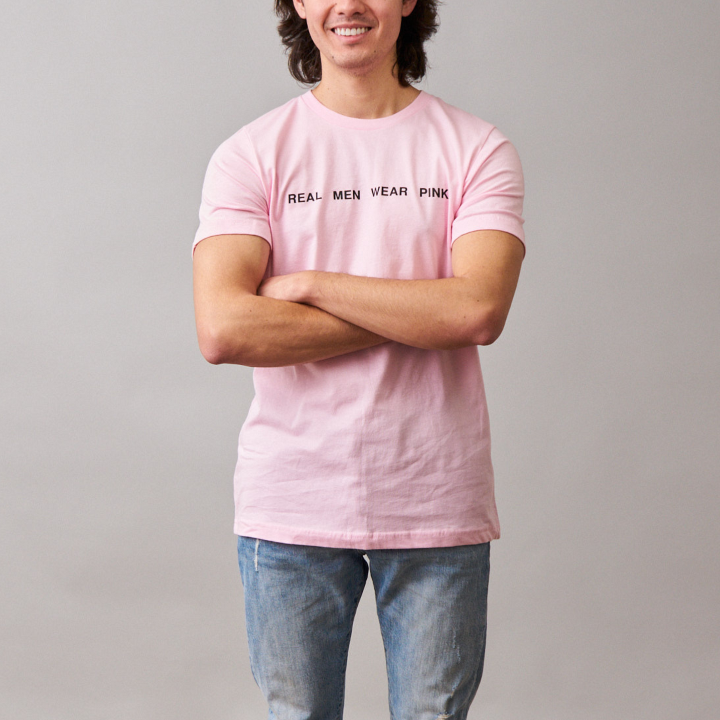 Man smiling wearing a pink t-shirt with 'REAL MEN WEAR PINK' printed on the front, paired with blue jeans